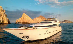 Luxury Yacht in Front of Cabo San Lucas Cliffs at Sunset A luxury yacht cruising near the rocky cliffs of Cabo San Lucas at sunset. The boat has a sleek design, large windows, and multiple decks, while the warm golden light from the sunset reflects off the yacht’s surface, enhancing its elegance.