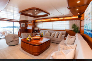 Elegant Yacht Living Room with Luxurious Wood Accents A spacious and stylish yacht living room featuring plush seating, a wooden coffee table, and large windows providing natural light. The space is decorated with vibrant flowers, cozy cushions, and soft lighting, offering an inviting and sophisticated atmosphere for relaxation. The design combines luxury with comfort, making it the ideal space to enjoy the views and unwind.