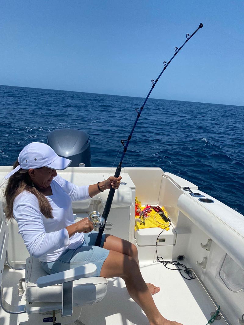 Fisherwoman engaged in sport fishing on a boat in Cabo San Lucas, holding a fishing rod while sitting in a captain's chair, with a clear ocean view.