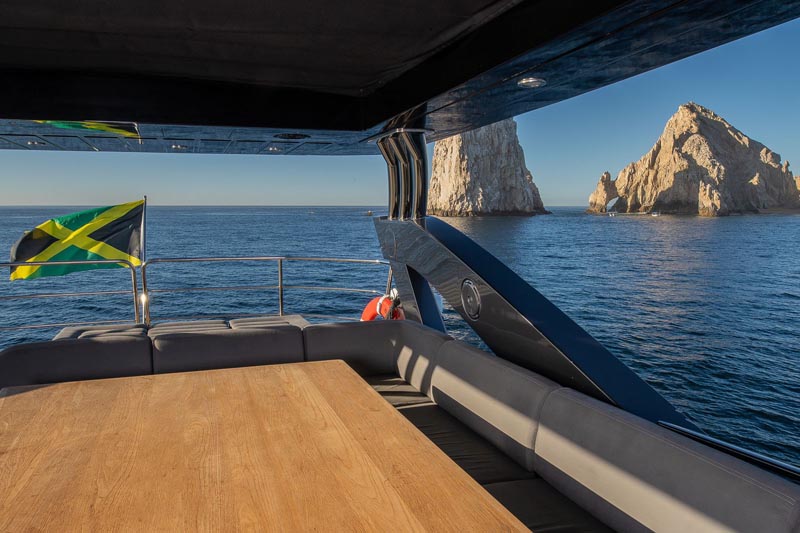 Stylish yacht deck with a wooden table and comfortable seating, overlooking Cabo San Lucas' famous arch and a Jamaican flag fluttering in the breeze.