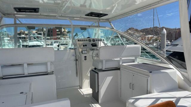 A clean, spacious cabin featuring a well-equipped cockpit of a boat. The white upholstery and open glass windows offer clear views of the harbor and nearby boats, with modern controls and storage areas seen on the dashboard. The bright sunlight enhances the boat's pristine look, highlighting its sleek design and the comfort provided by the open, airy space. The image captures a perfect moment of leisure and luxury on the water.