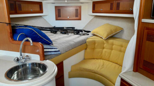 Cabo San Lucas Fishing Charters An interior view of the "Game On" boat's cozy cabin area, showcasing a comfortable bed with neatly arranged pillows. The surrounding space features a circular cushioned seating area with yellow upholstery, offering a relaxing spot for passengers. A small kitchenette sink adds functionality, while fishing equipment rests on the bed, emphasizing the boat's dual-purpose for both leisure and adventure. The warm wood finishes and well-designed layout provide a welcoming and efficient use of space for guests.